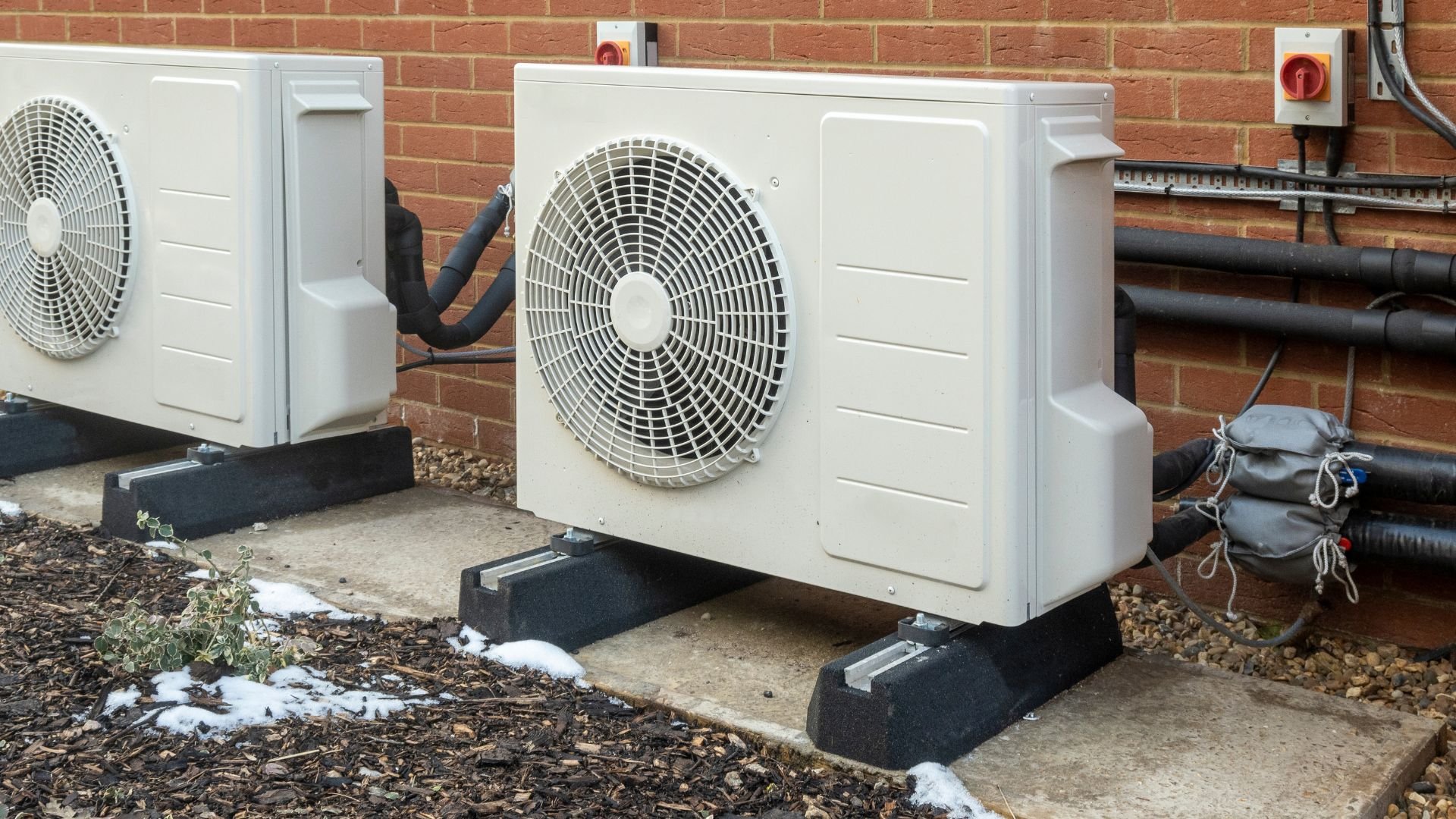 Two white air conditioning units mounted on brick wall with snow nearby